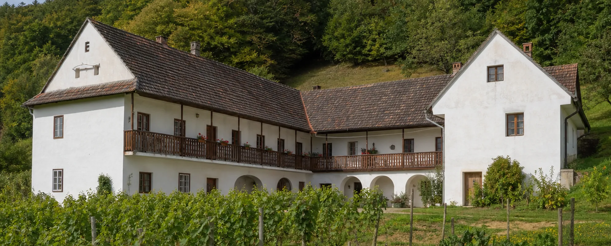 Weiß verputzter Vierkanthof mit Holzbalkon, davor ein kleiner Weingarten